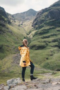 Blonde Girl in a yellow raincoat standing in front of some breathtaking mountains.