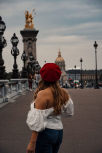 Weekend in Paris - Pont Alexandre III. I am wearing a white shoulder free blouse and a red beret, a perfect outfit for Paris. In the background is the dome of Invalides and the golden details of the bridge.