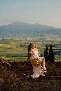 Woman sitting on a wall in a white sun dress. In the background are the green hils of Tuscany.