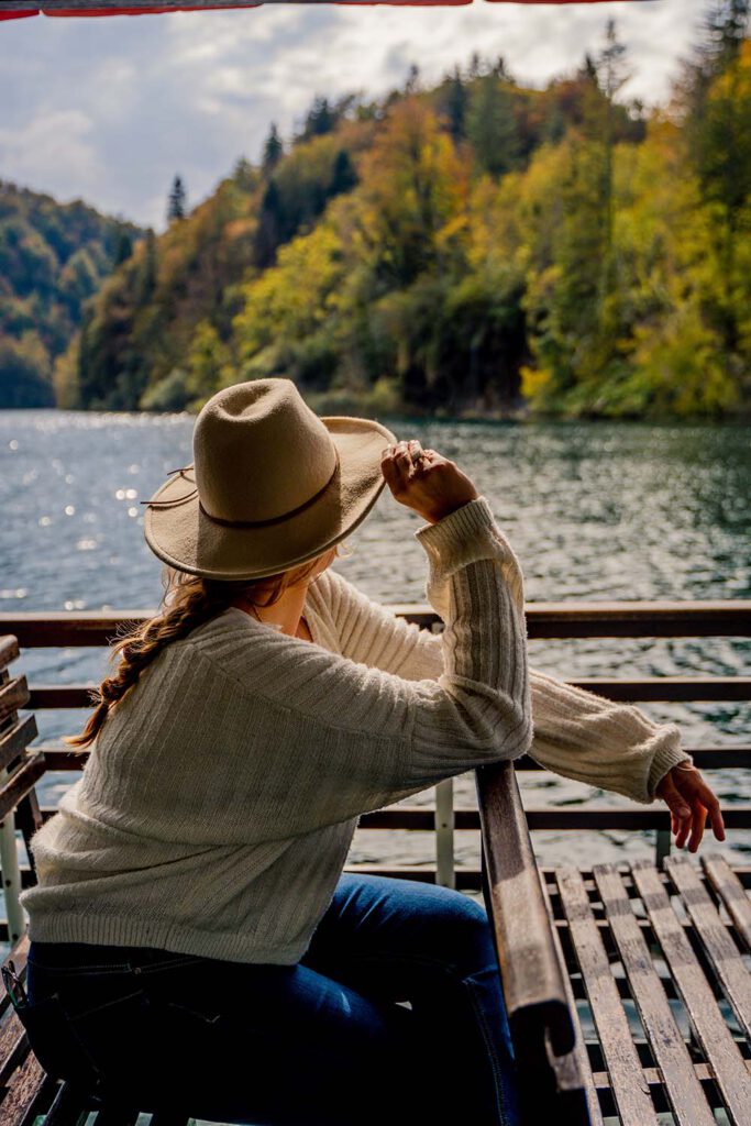 Paula from La Vie En Marine sits on a ferry in a cozy sweater overlooking the Plitvicer Lakes, which is framed by colorful leaves during autumn in Europe!