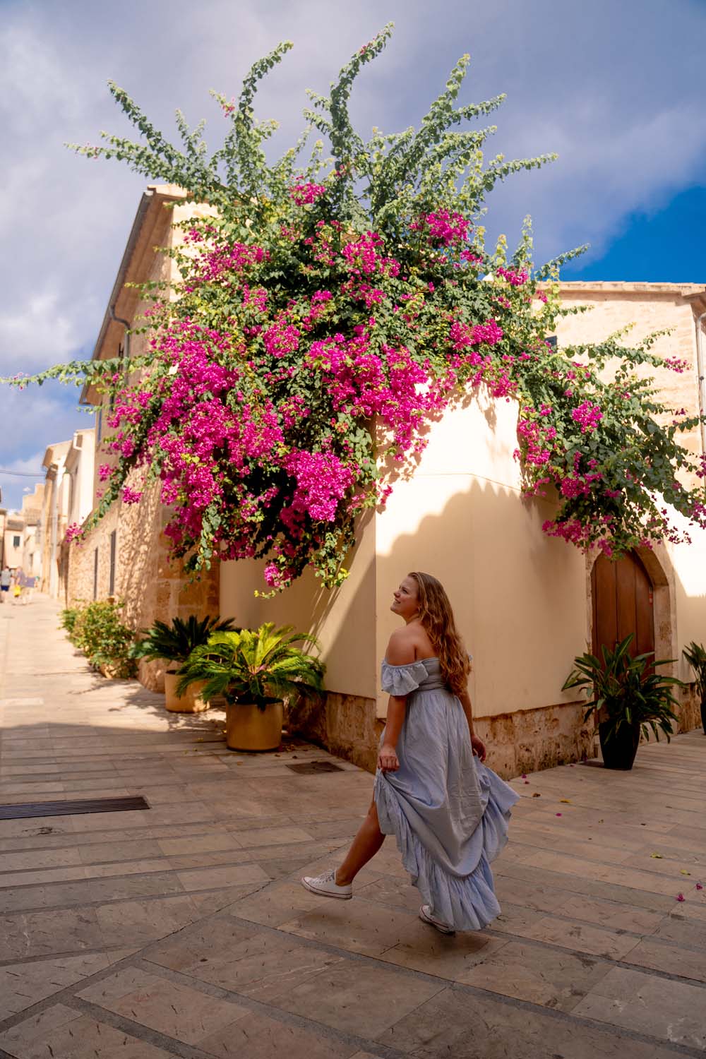 A blonde girl stands in front of beiges houses that are covered in green plants with lush pink flowers in Alcúdia, Mallorca.