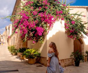 A blonde girl stands in front of beiges houses that are covered in green plants with lush pink flowers in Alcúdia, Mallorca.