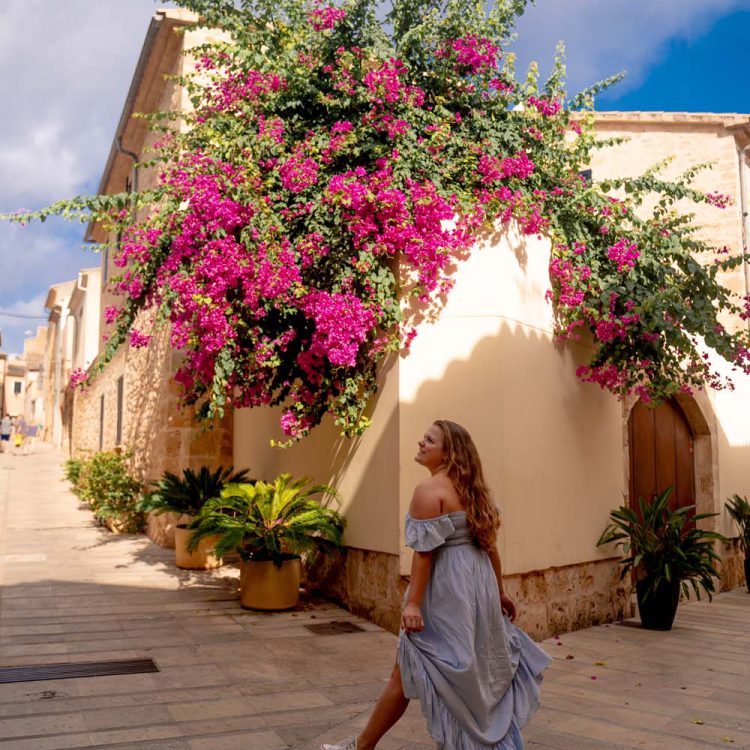 A blonde girl stands in front of beiges houses that are covered in green plants with lush pink flowers in Alcúdia, Mallorca.